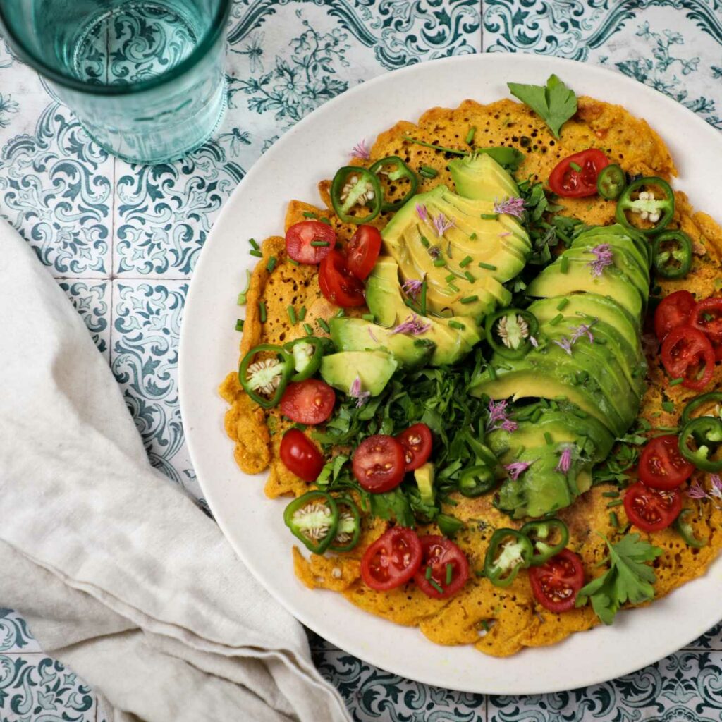 Veganská omeleta je zobrazena s hlávkovým salátem a avokádem na talíři na barevných dlaždicích.