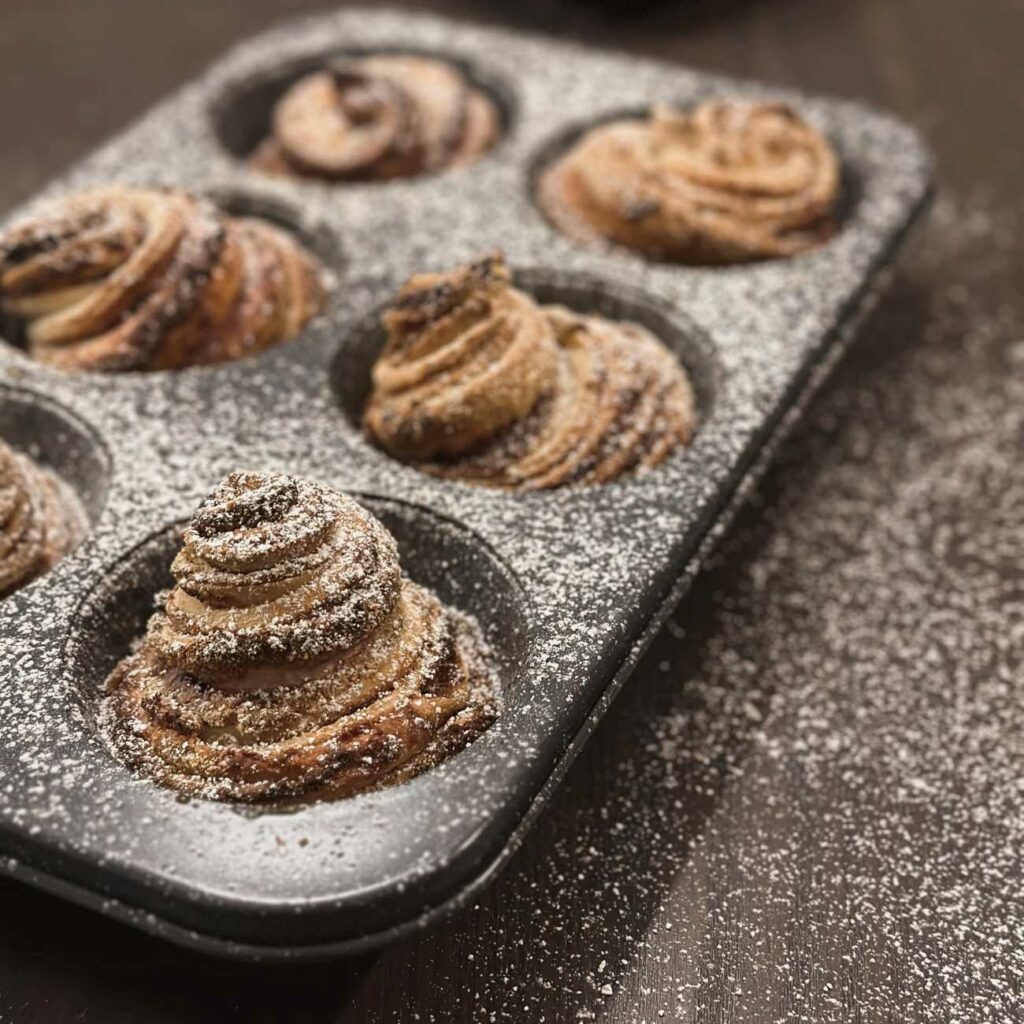 Cruffins werden in einer Muffinform mit Puderzucker bestreut gezeigt.