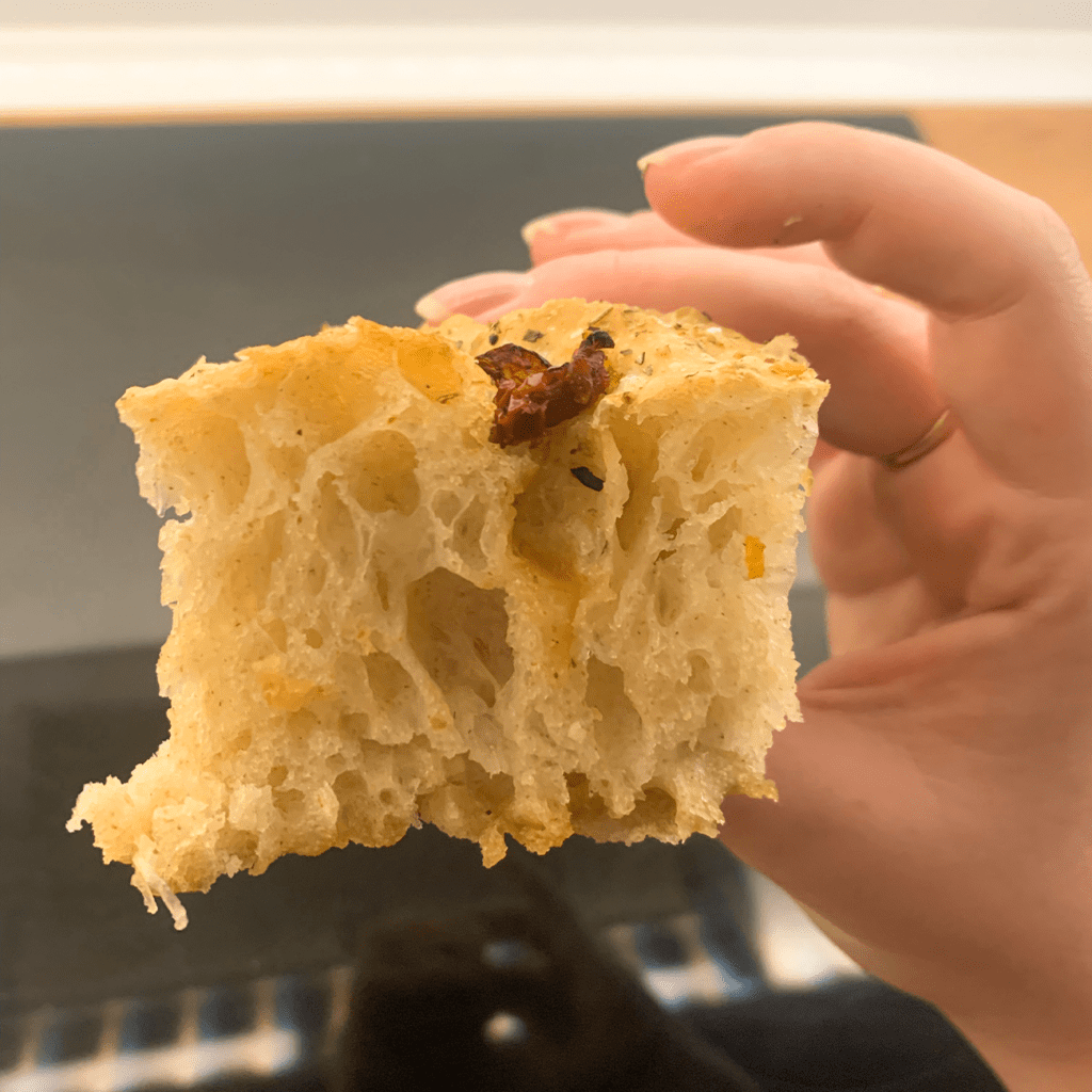 Focaccia wird angeschnitten in die Kamera gehalten, sodass viele Bläschen und ein lockerer Teig zu sehen sind.